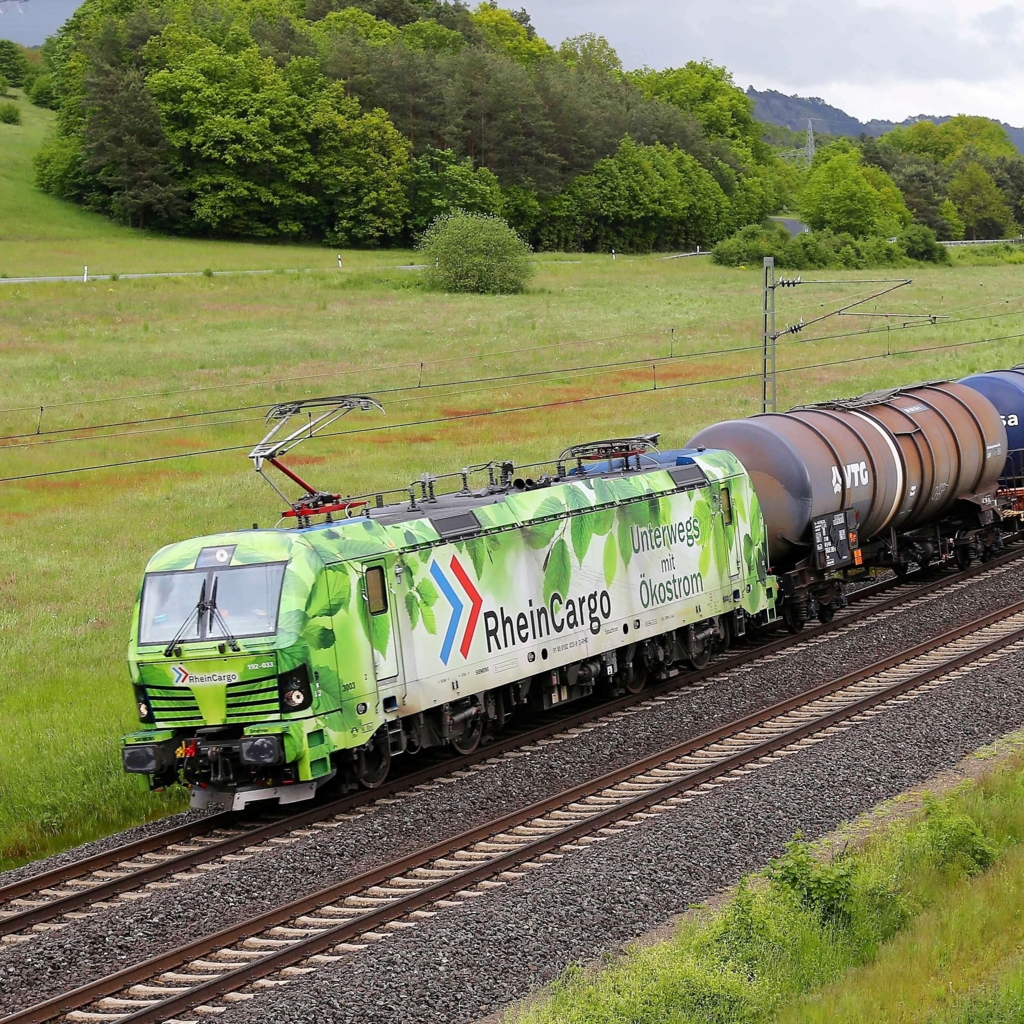 Foto eines RheinCargo-Güterzuges, der durch grüne Landschaft fährt