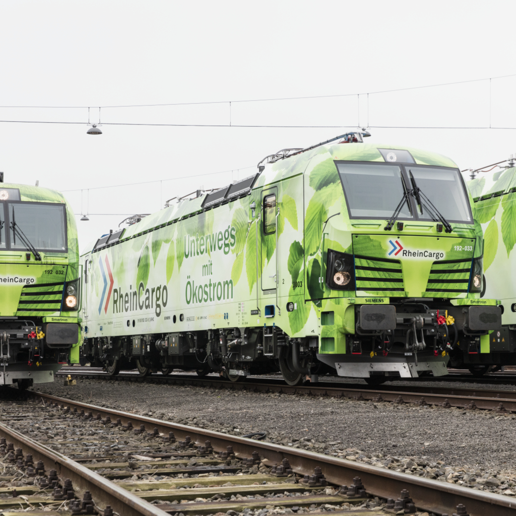 Drei grün folierte Elektrolokomotiven von RheinCargo mit der Aufschrift „Unterwegs mit Ökostrom“ stehen nebeneinander auf Gleisen eines Rangierbahnhofs. Die Lokomotiven sind mit Blattmotiven verziert und symbolisieren umweltfreundlichen Güterverkehr.
