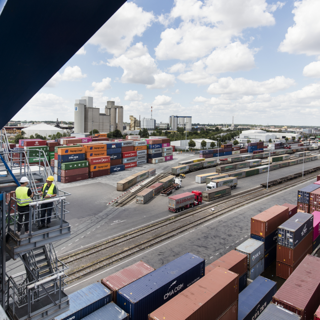 Blick auf ein Containerterminal mit gestapelten Überseecontainern, Eisenbahnschienen, LKWs und zwei Personen in Sicherheitskleidung auf einer Metallplattform im Vordergrund. Im Hintergrund sind Industrieanlagen und Lagerhallen zu sehen, unter einem leicht bewölkten Himmel.