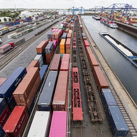 Luftaufnahme eines Containerterminals mit Güterzügen, Lkw und Frachtschiffen an einem Binnenhafen mit Kränen und Lagerflächen.