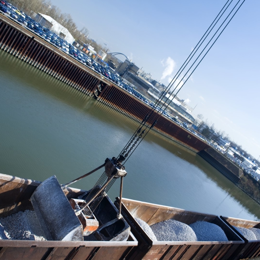 Schüttgut-Umschlag am Hafen: Ein Kran befüllt offene Güterwaggons mit Schotter oder Kies, während im Hintergrund Wasserfläche, Lagerflächen und Industriegebäude zu sehen sind.
