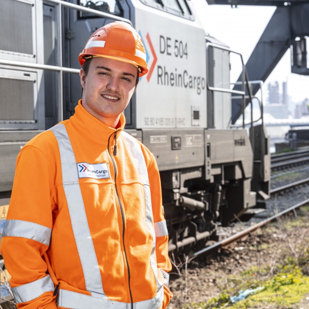 Ein Mitarbeiter von RheinCargo steht in orangefarbener Schutzkleidung und Helm vor einer Lokomotive mit der Aufschrift „DE 504 RheinCargo“. Im Hintergrund sind Gleise, ein Verladekran und Teile eines Hafens zu sehen. Die Aufnahme entstand bei sonnigem Wetter und vermittelt den Eindruck moderner, sicherer Logistikarbeit im Bahnbereich.