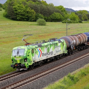 Eine grüne Elektrolokomotive der RheinCargo mit der Aufschrift „Unterwegs mit Ökostrom“ zieht einen langen Zug aus dunklen Kesselwagen über eine zweigleisige Bahnstrecke durch eine hügelige, grüne Landschaft mit Wiesen, Bäumen und bewölktem Himmel im Hintergrund.