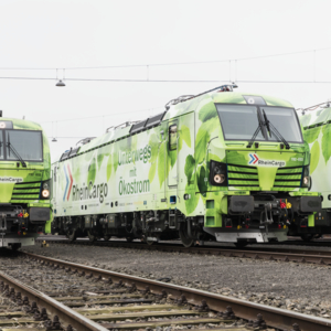 Drei grün folierte Elektrolokomotiven von RheinCargo mit der Aufschrift „Unterwegs mit Ökostrom“ stehen nebeneinander auf Gleisen eines Rangierbahnhofs. Die Lokomotiven sind mit Blattmotiven verziert und symbolisieren umweltfreundlichen Güterverkehr.