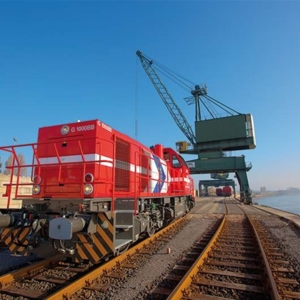 Eine rote Lokomotive der RheinCargo steht auf Gleisen am Rheinufer. Im Hintergrund befindet sich ein großer grüner Hafenkran, daneben verlaufen Schienen und Wasser. Der Himmel ist klar und blau, die Szene zeigt den Hafenbetrieb bei sonnigem Wetter.