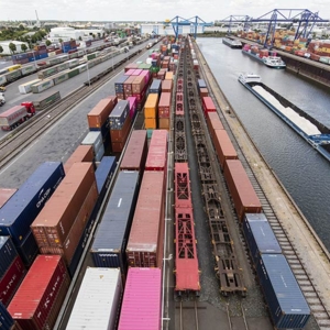 Luftaufnahme eines Containerterminals mit Güterzügen, Lkw und Frachtschiffen an einem Binnenhafen mit Kränen und Lagerflächen.