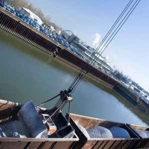 Schüttgut-Umschlag am Hafen: Ein Kran befüllt offene Güterwaggons mit Schotter oder Kies, während im Hintergrund Wasserfläche, Lagerflächen und Industriegebäude zu sehen sind.